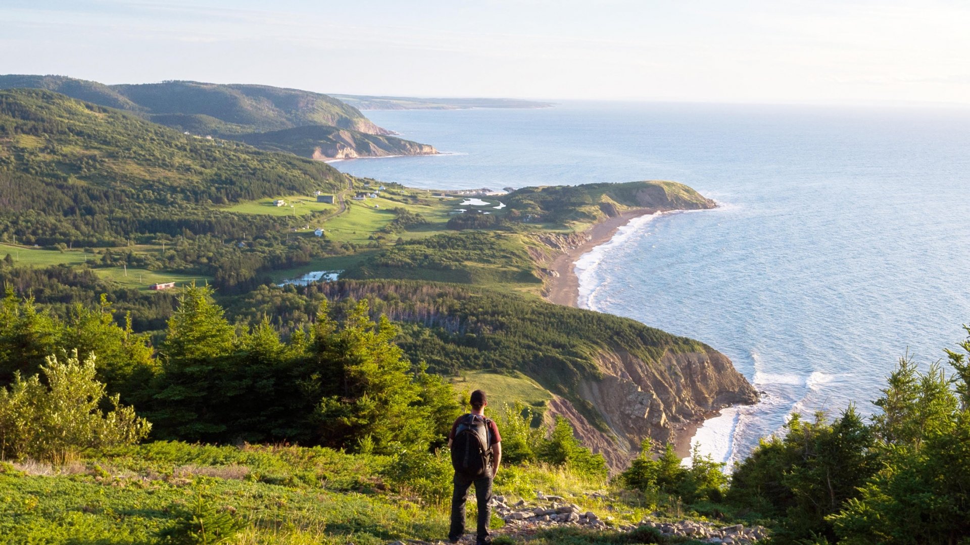 A hiker stands on a grassy hill overlooking the ocean