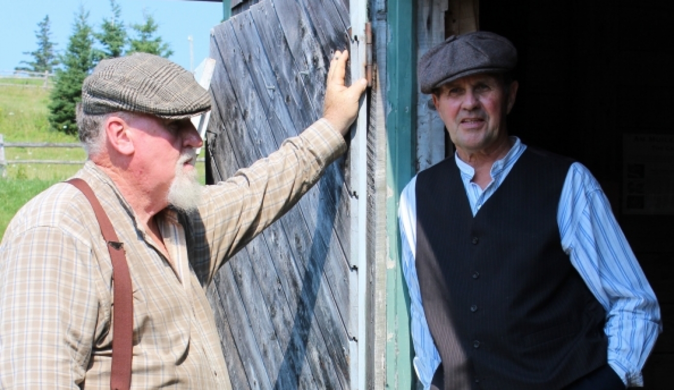 two older men in flat caps talk near a barn